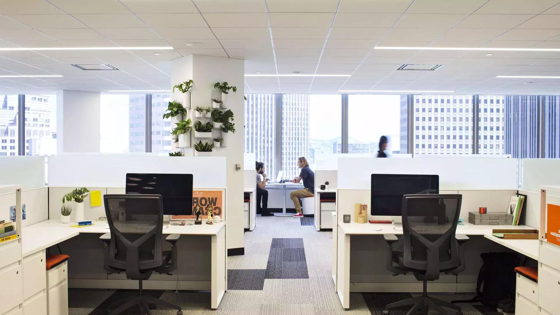 Scène d'intérieur dans un immeuble de bureaux à San Francisco, avec des bureaux au premier plan et deux personnes travaillant à une table devant de grandes fenêtres.