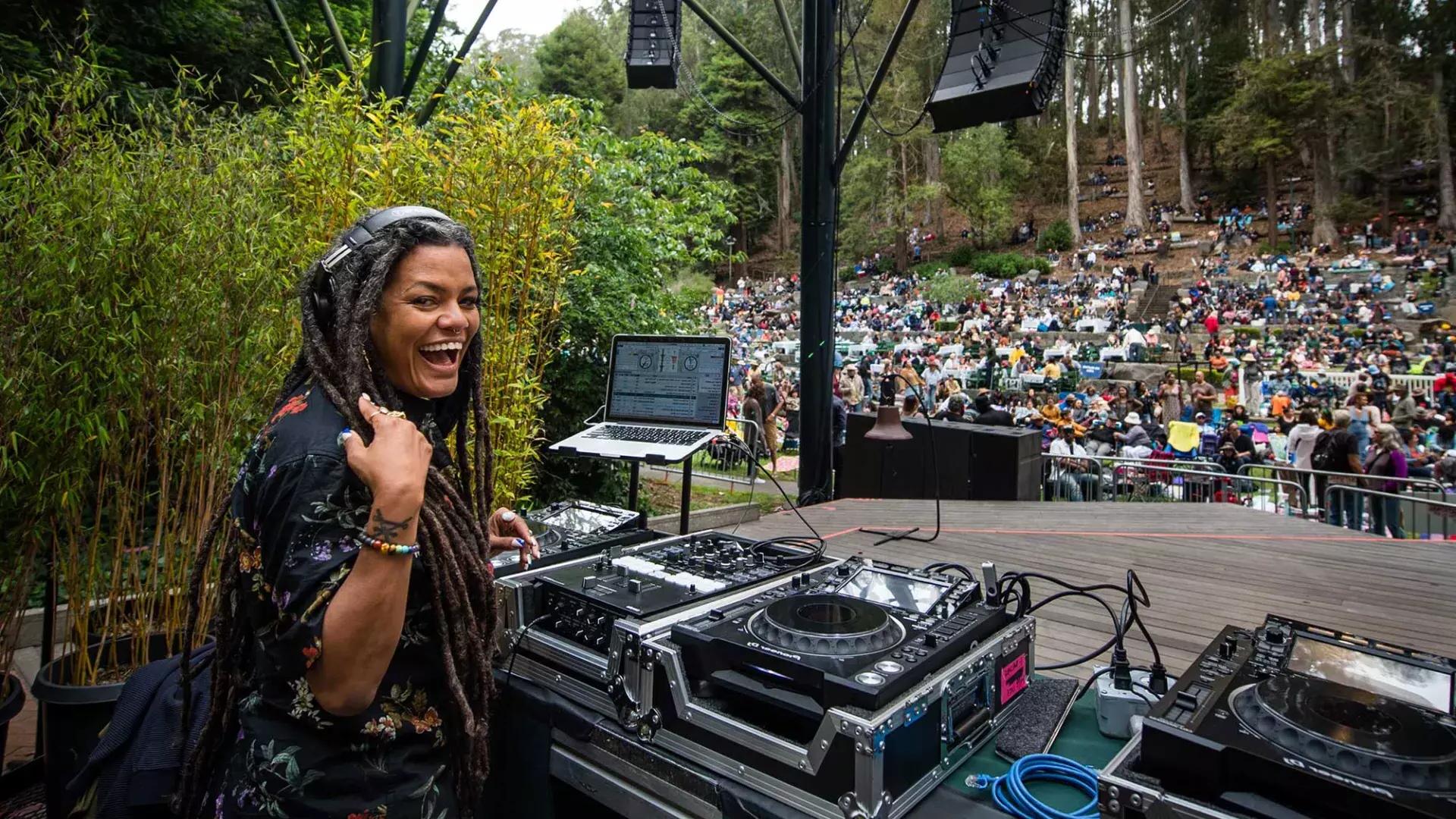 斯特恩格罗夫 페스티벌(Stern Grove Festival)에서 DJ를 하고 있는 한 여성이 어깨 너머로 카메라를 바라보며 미소를 짓고 있습니다.
