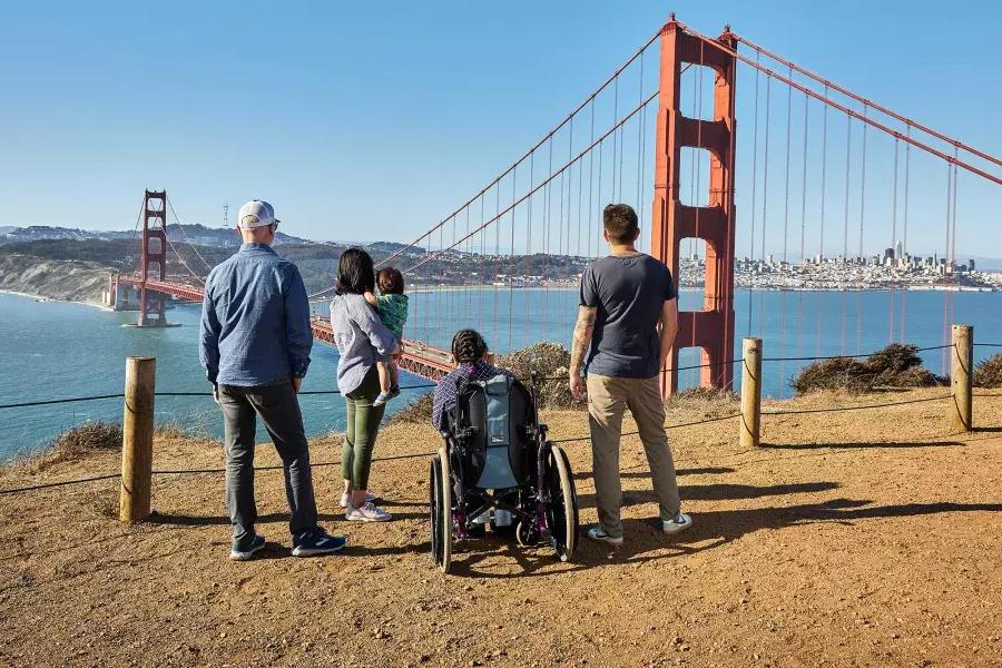 A group of people, including one person in a wheelchair, is seen from behind as they look at the Golden Gate Bridge from the Marin Headlands.