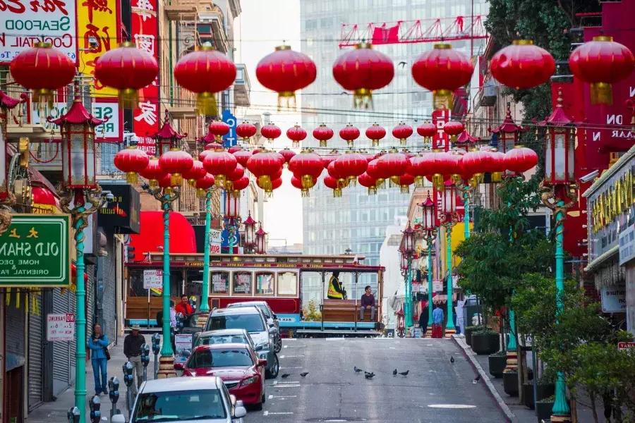 Una strada collinare nella Chinatown di San Francisco è raffigurata con lanterne rosse penzolanti e un tram che passa.