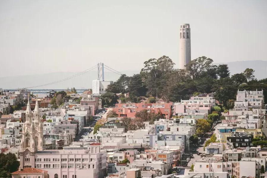 La Coit Tower di San Francisco è raffigurata con il Bay Bridge sullo sfondo e una collina ricoperta di case in primo piano.