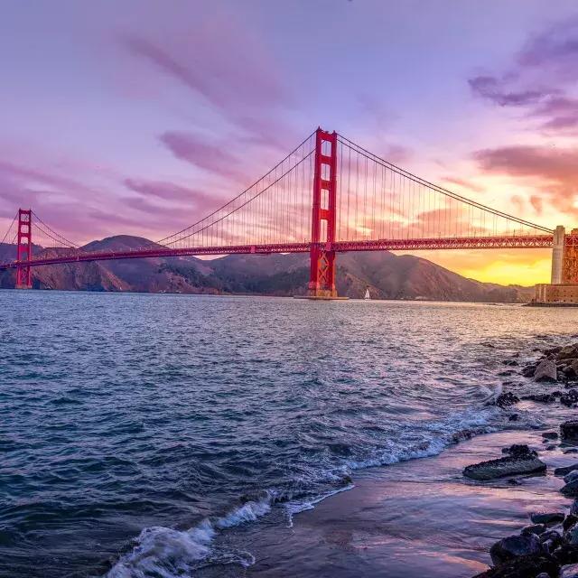 Il Golden Gate Bridge al tramonto con un cielo multicolore e la Baia di San Francisco in primo piano.