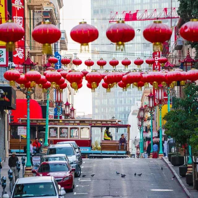 Abgebildet ist eine hügelige Straße in San Franciscos Chinatown mit baumelnden roten Laternen und einer vorbeifahrenden Straßenbahn.