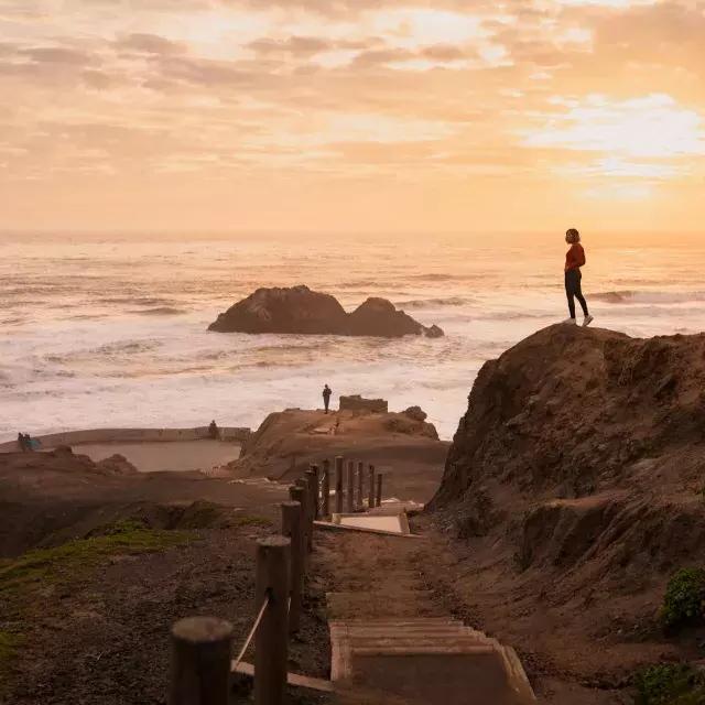 Duas pessoas estão sobre rochas com vista para o oceano em Sutro Baths, em São Francisco.